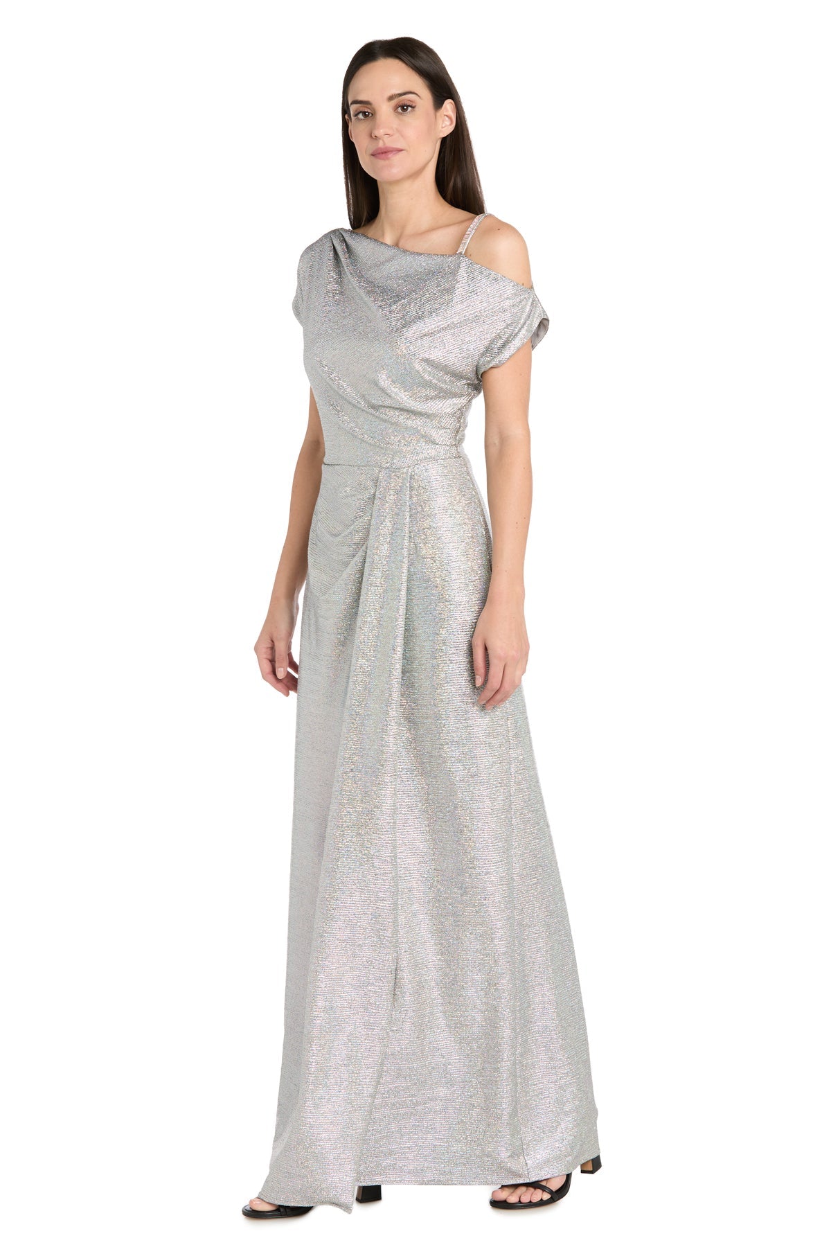 A woman wears the R&M Richards 3758 Silver Metallic Formal Dress, featuring a floor-length design with off-shoulder draped detailing, paired with black heeled sandals, standing against a white background.